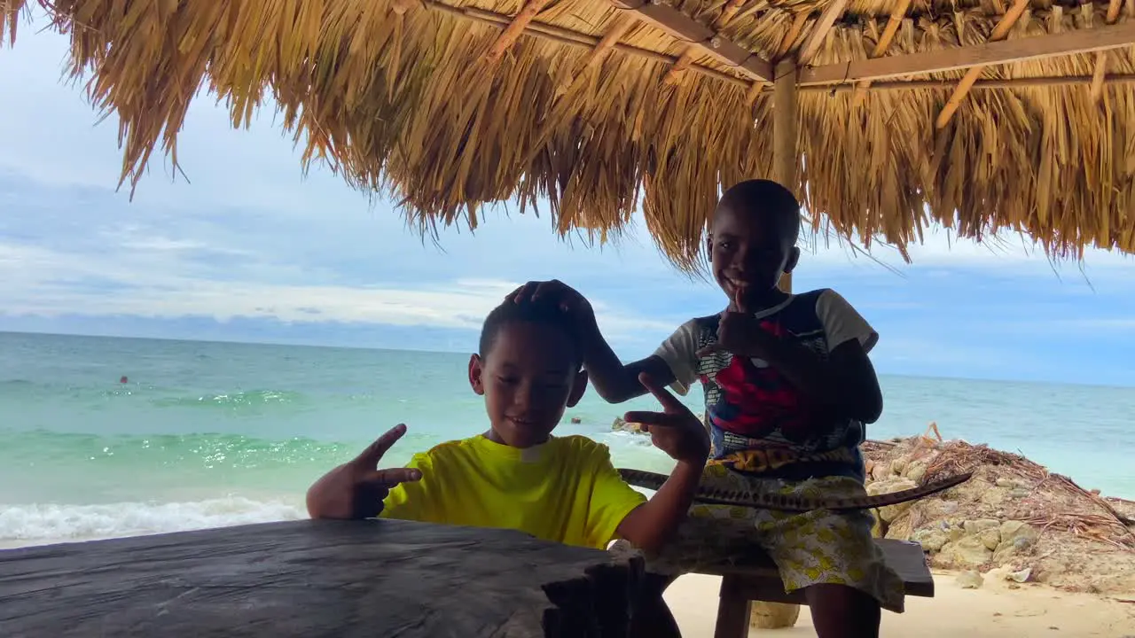 Two black kids posing and smiling to camera under a thatched roof on a tropical beach