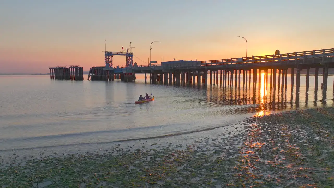 Young couple take off and paddle together in sea kayak at sunrise floating under pier with sun glare reflecting off bay water near seattle washington aerial drone view