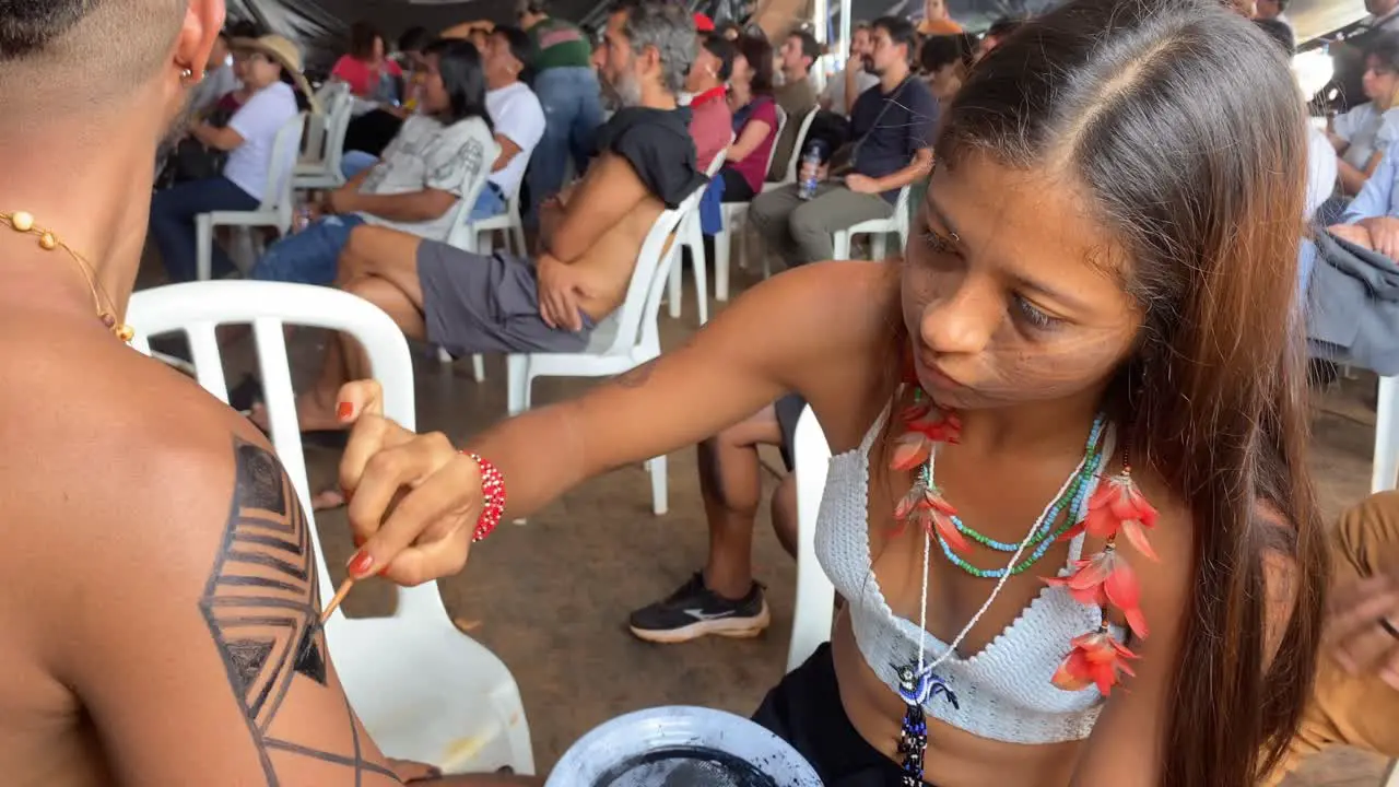 A beautiful native amazon woman with red colored plumes in her hair makes traditional body painting on a man