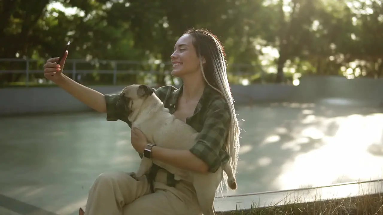 Cheerful Girl Taking Selfie Photo With Cute Pug Puppy In Green City Park Holding Smartphone In Summer Day