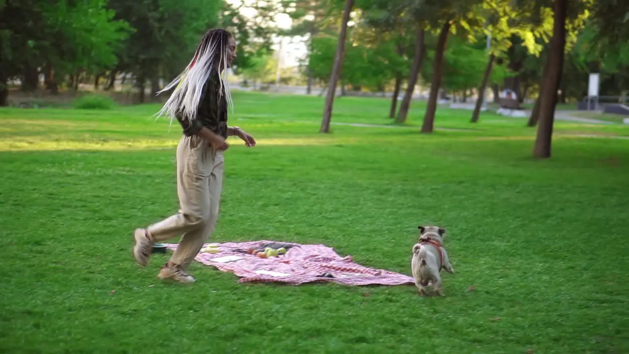 Happy Young Woman Having Fun With Her Dog Outdoors Running Chasing It Around Plaid With Picnic On Grass