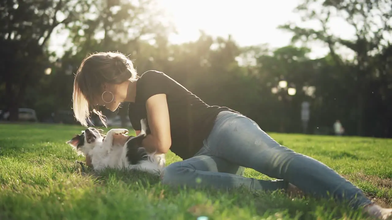 Front Footage Of A Woman In Jeans And Black T Shirt Lying On The Green Grass In The Park And Playing With Her Pet