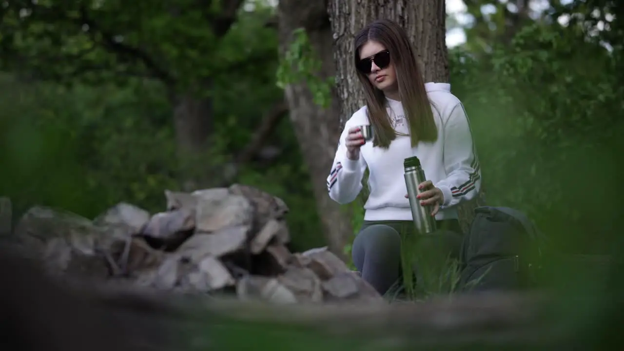 Young caucasian girl sitting in forest pouring herself hot drink from thermos bottle and drinking it