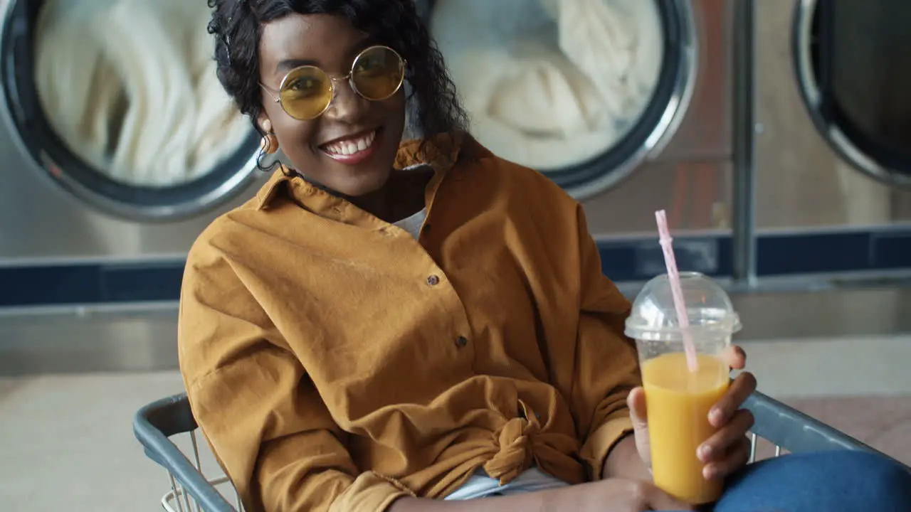 Pretty And Happy Girl Sitting In Trolley And Drinking Orange Juice With Straw Resting And Waiting For Clothes To Be Washed