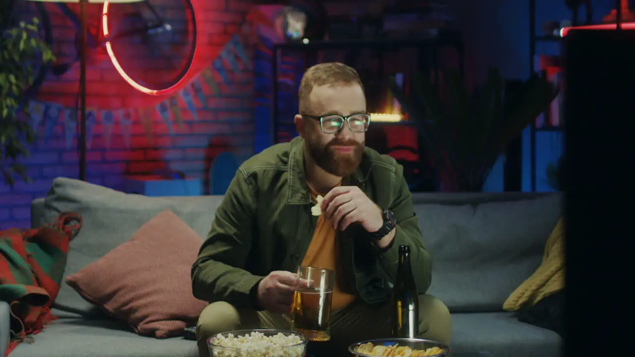 Young Guy In Glasses Sitting On The Couch With Beer And Snacks An Worrying About Sport Match On Tv At Night