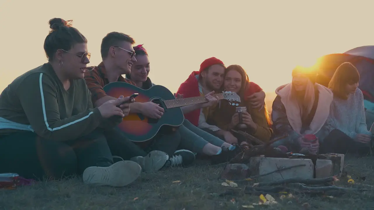 young people sit with guitar at burning bonfire in evening