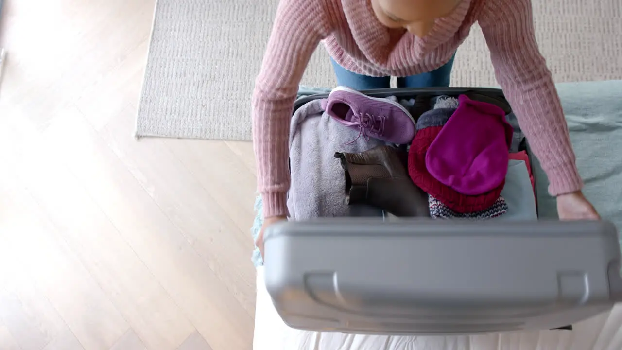 Overhead view of biracial woman packing suitcase in sunny bedroom slow motion