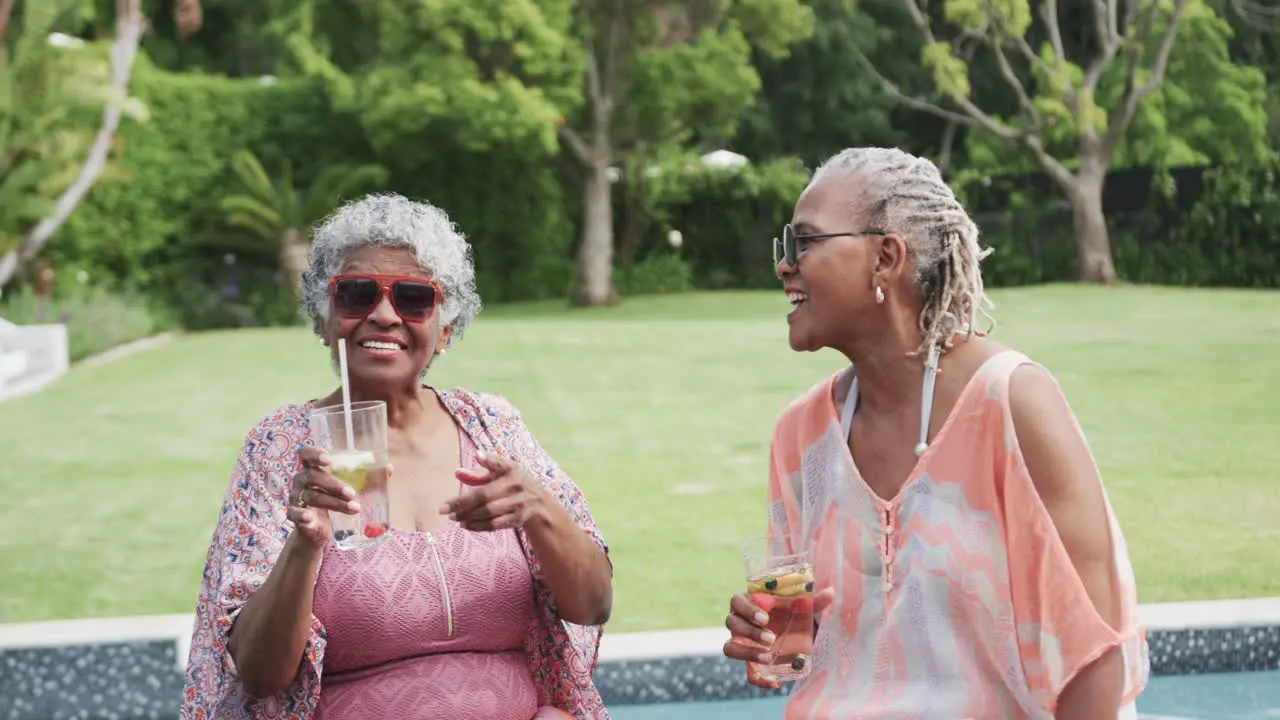 Happy senior african american female friends with sunglasses and drinks sitting by pool slow motion