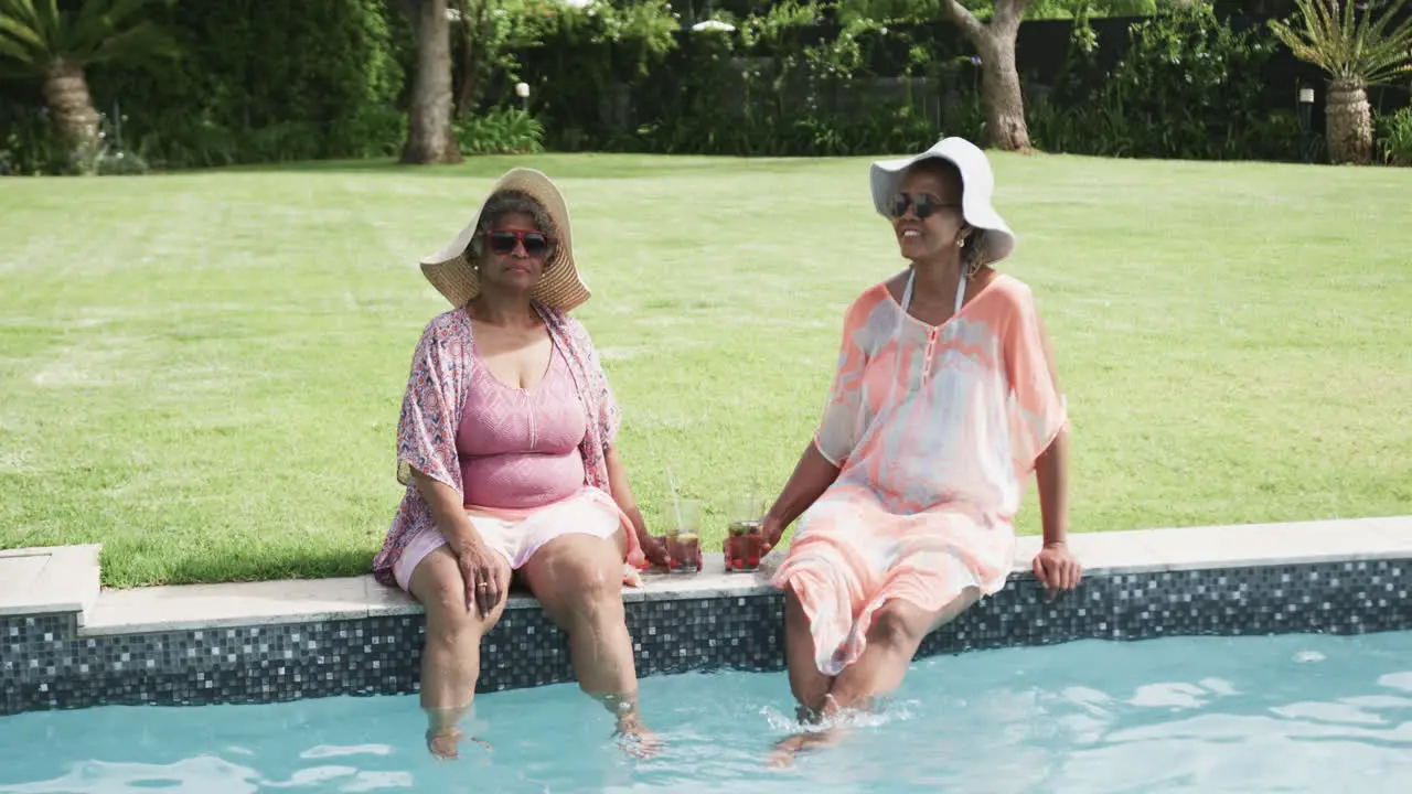 Happy senior african american female friends in sunhats sitting by pool with drinks slow motion