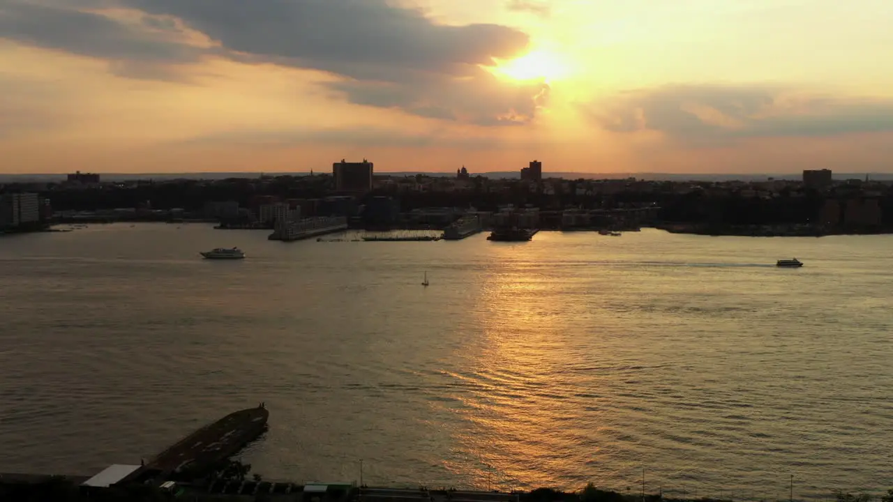 Aerial footage of the Hudson River in New York as the drone rises focuses on the sunset while two boats go by in opposite directions with a sailboat in the middle