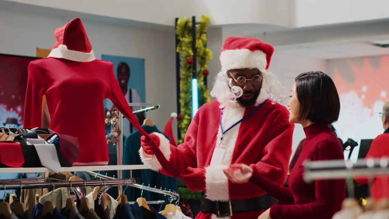 African american employee dressed as Santa Claus helping client pick festive outfit in Christmas shopping store during winter holiday season Worker assisting customer in fashion shop