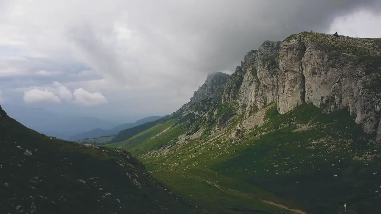 Drone backward shot revealing a mountain ridge with low clouds and an alpine valley with green grass