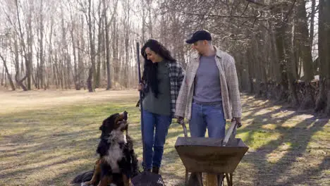 Cucasian couple and their dog holding a wheelbarrow and a rake in the countryside