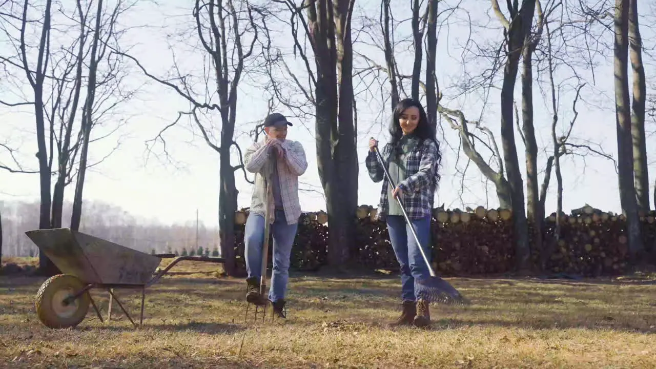 Caucasian woman removing weeds with a rake in the field and talking with her partner The man is standing near to her while holding other rake