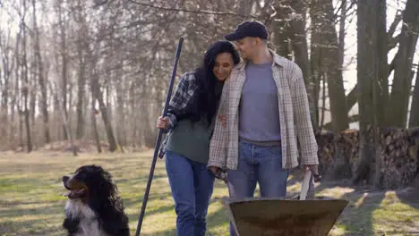 Caucasian couple and their dog holding a wheelbarrow and a rake in the countryside