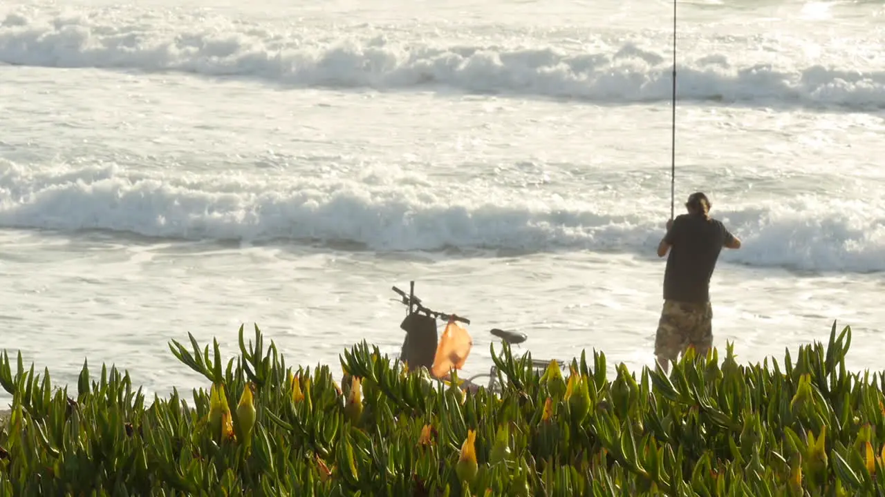 Man in Hat Casts Fishing Line into the Ocean as Waves Crash Plants in Foreground