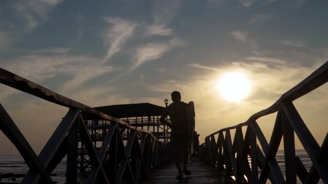 A slow motion tracking shot of a male surfer jogging down the pier at dusk