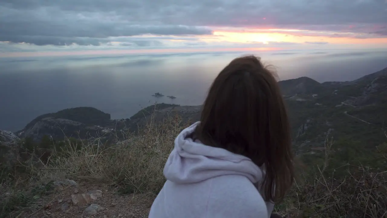 Girl squat on a high cliff and watch the sunset through cloudy sky over Adriatic sea