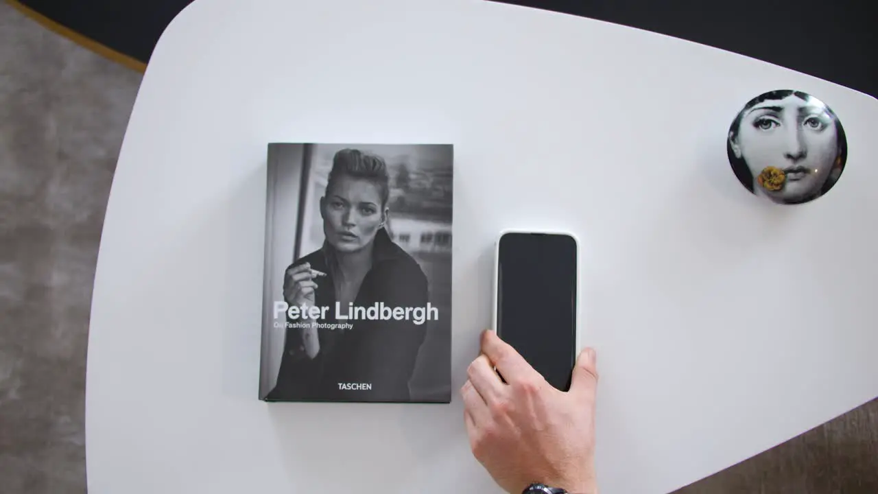 view from above of a small white designer coffee table on which a book with a woman in black and white on the cover a human hand places a smartphone beside it blurred gray floor