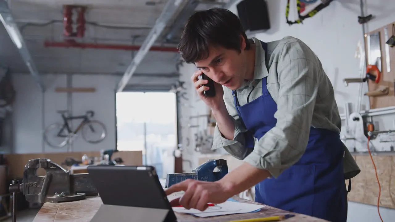 Male Business Owner In Workshop Using Digital Tablet And Making Call On Mobile Phone