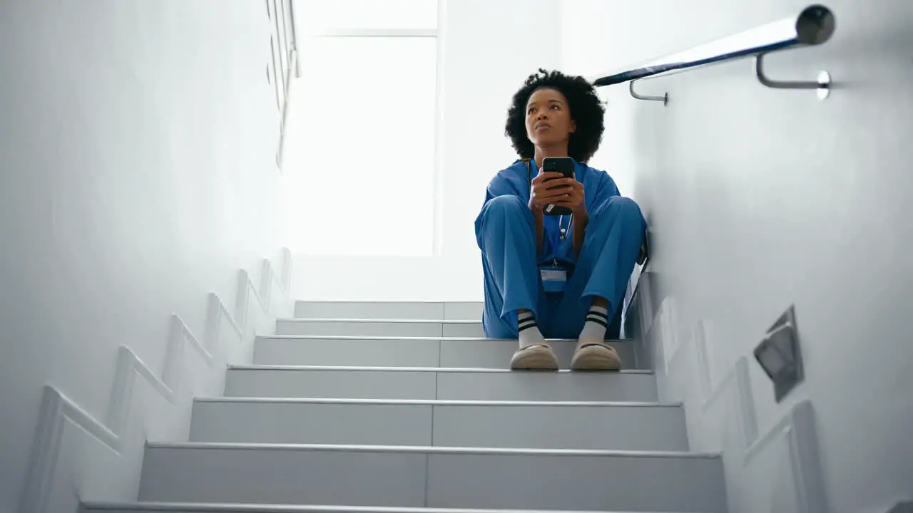 Stressed Female Nurse Wearing Scrubs Browsing On Mobile Phone Sitting On Stairs In Hospital