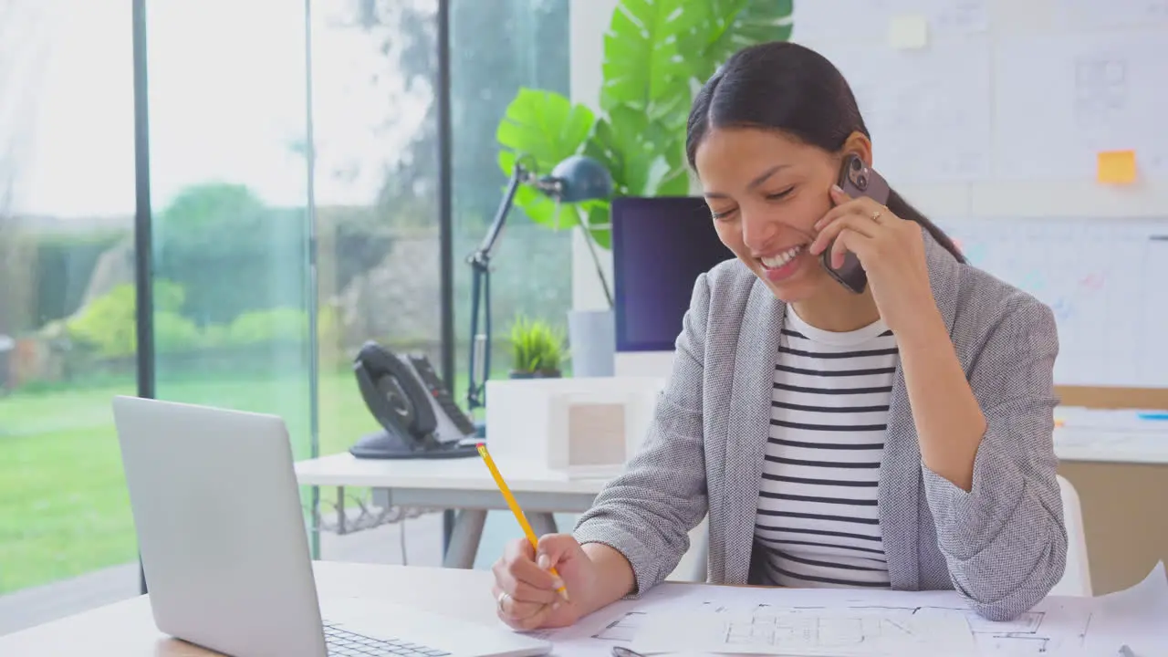 Female Architect Working In Office Sitting At Desk Talking On Mobile Phone