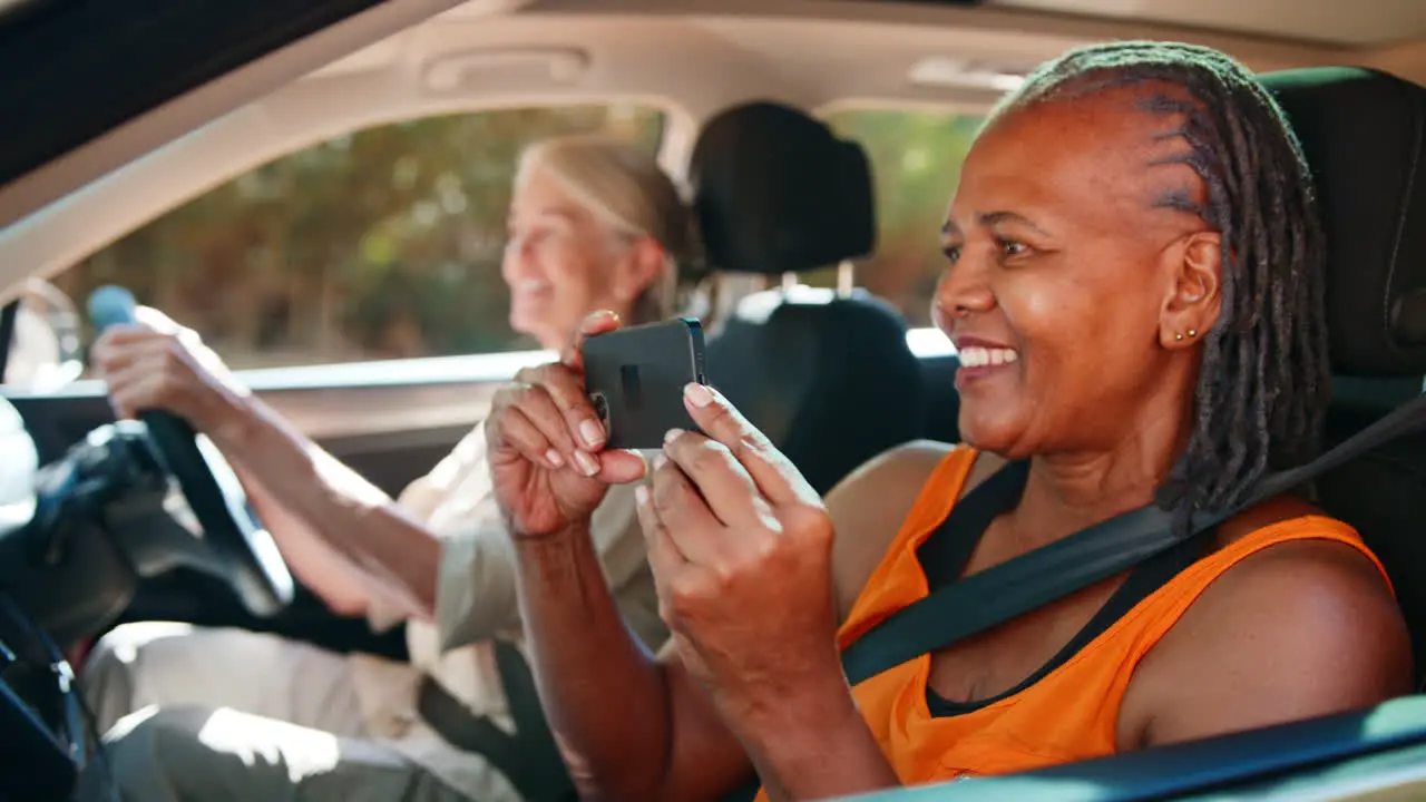 Passenger Taking Photo On Mobile Phone As Two Senior Female Friends Enjoy Day Trip Out In Car