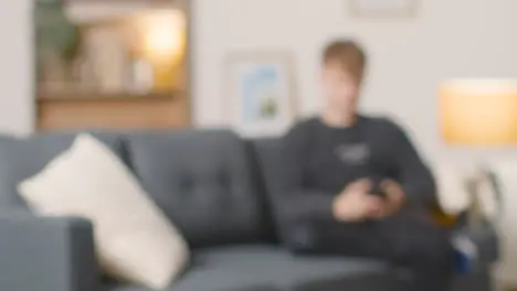 Defocused Shot Of Young Man Sitting On Sofa At Home Using Mobile Phone