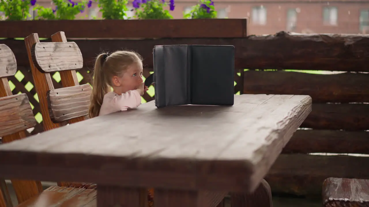 Little girl with tablet sits and waits rain end in cafe