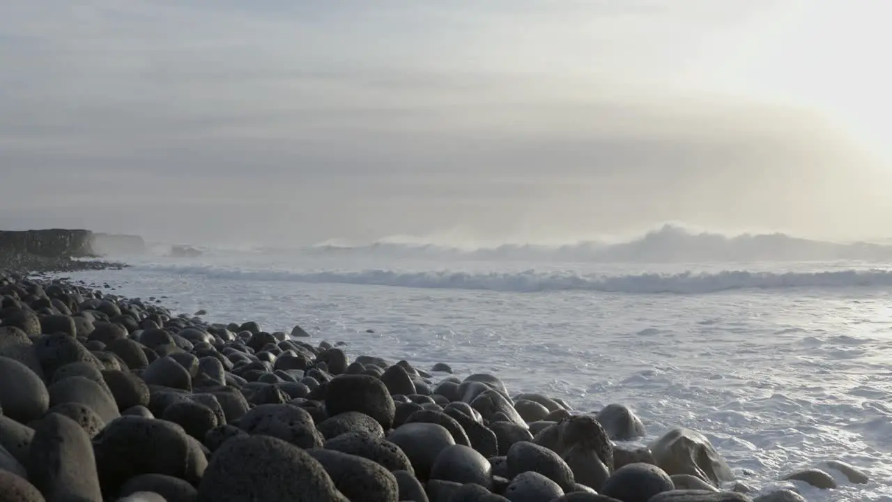 Hazy ocean shore view in Iceland with waves spraying mist bright sunshine rounded stones on beach