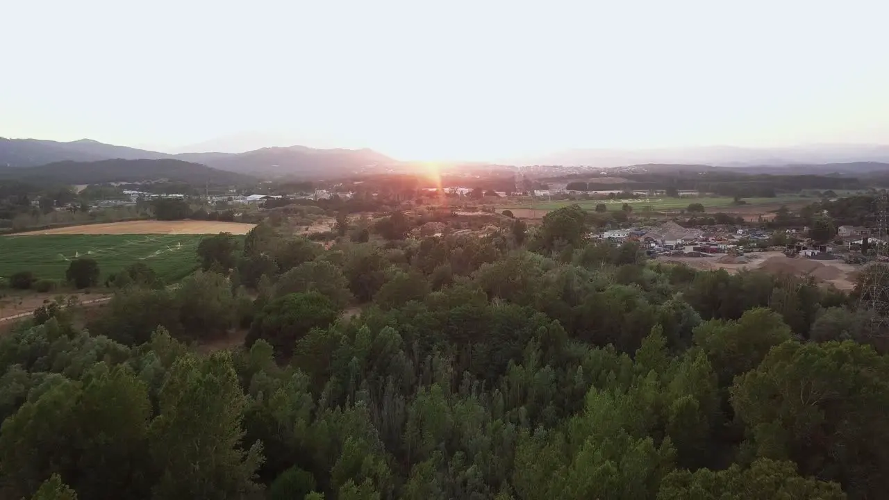 Aerial drone shot flying over a forest with fields on the left and a town on the right an a magnificent sunset over the mountains in the background