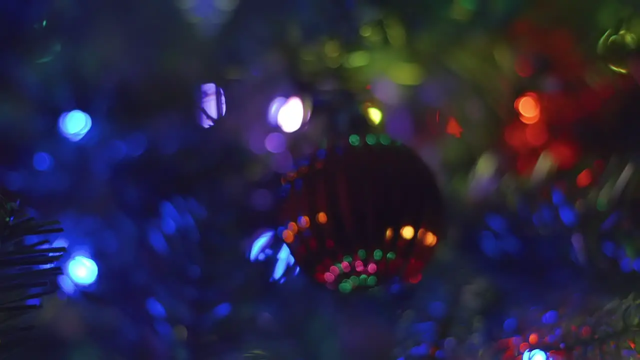 Panning and zooming away from red and textured bulb ornament with steady multi-colored bokeh lights on a Christmas tree