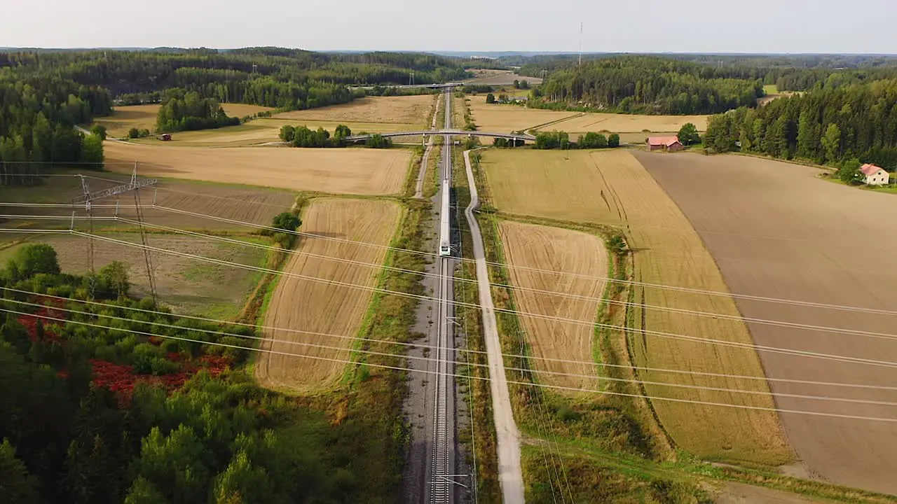 High altitude tracking aerial drone view of high speed passenger train traveling on long straight track passing under multilane highway bridge with fields and forests on both sides