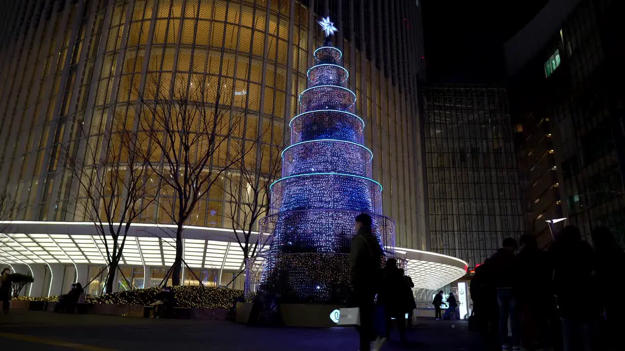 Decorated Christmas tree with colorful lights outside the Lotte World Tower with last-minute shoppers walking by on the eve of the holiday at night