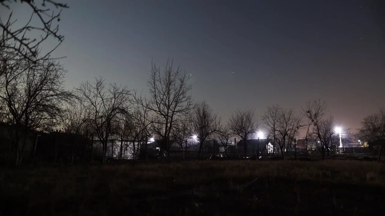 View Of Houses Surrounded By Leafless Trees With Flashes Of Lightning Over The Grass Field In The Countryside At Night