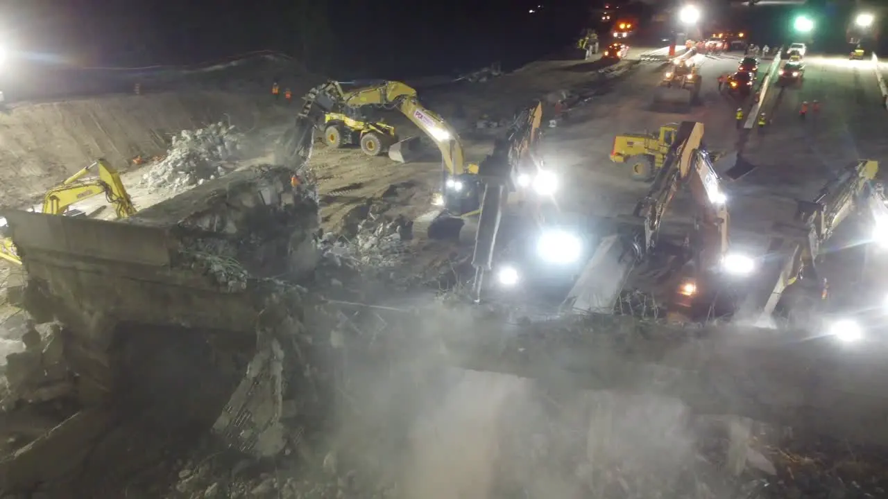 large group of heavy equipment demolishes a heavily reinforced concrete overpass in the night overhead drone showcases dusty night environment