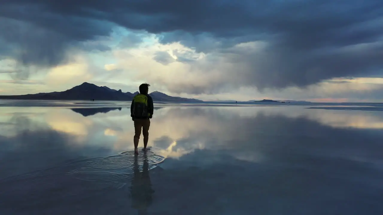 Silhouette of Man With Backpack Walking in Surreal Landscape of Salt Flats With Stormy Clouds and Sky Mirror Reflection on Water Cinematic Approaching View