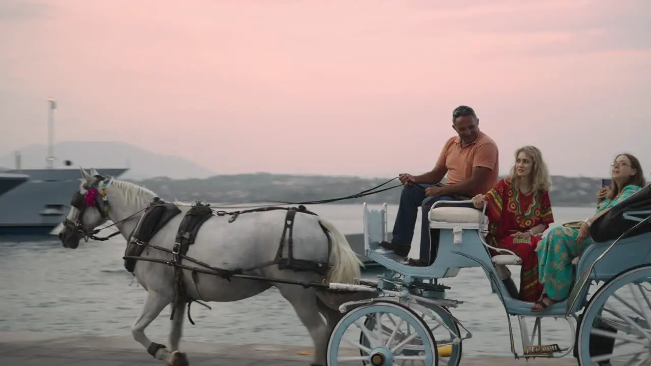 A light blue traditional carriage with a withe horse and tourists riding by in front of the Grand Poseidonion Hotel at romantic red sunset in the evening of spetses coastal harbour pier