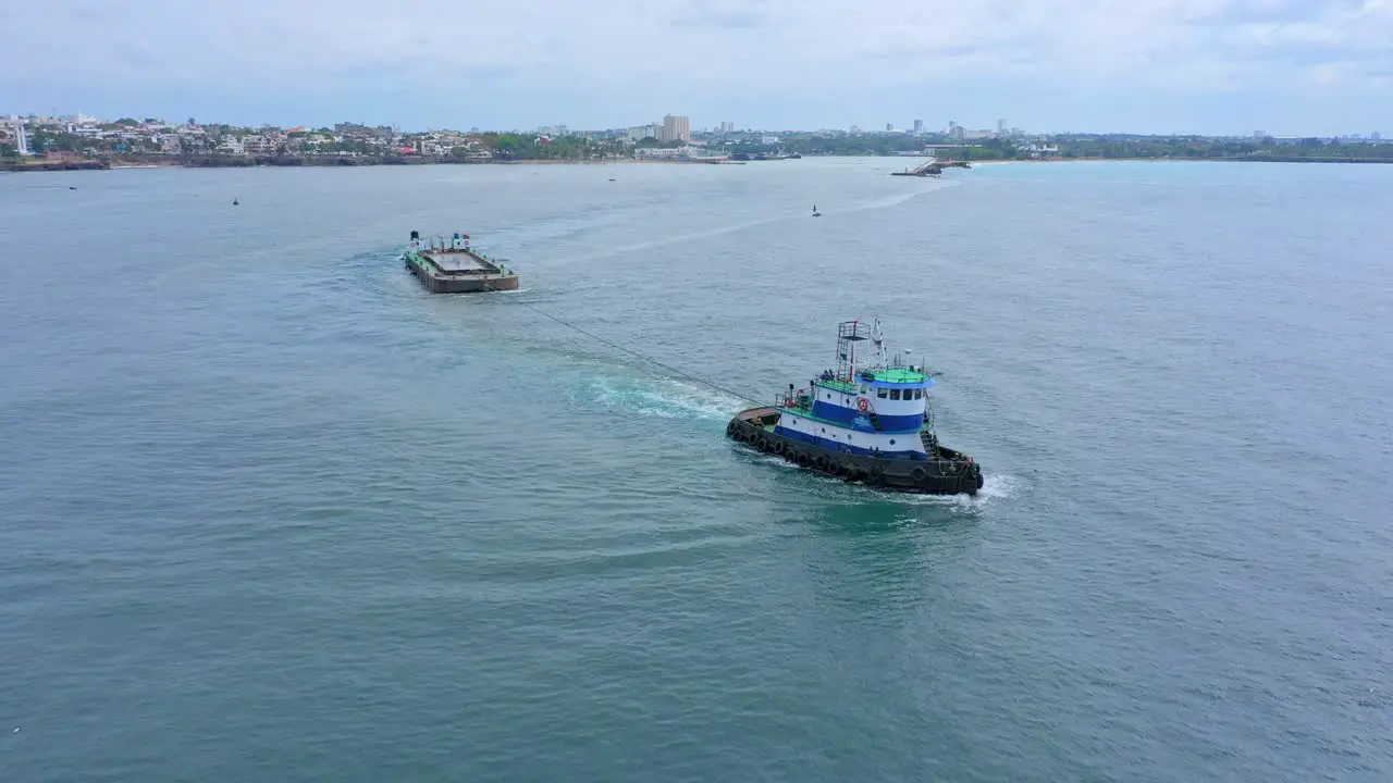 Tugboat drags platform out of harbor in open sea with city in background