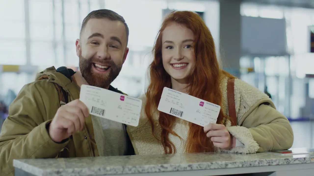 Portrait Of The Young Cheerful Couple Standing At The Checking Desk At The Airport With Tickets In Hands And Smiling To The Camera