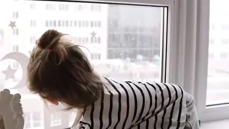 A little girl is sitting on the windowsill the cat is looking at her from the floor