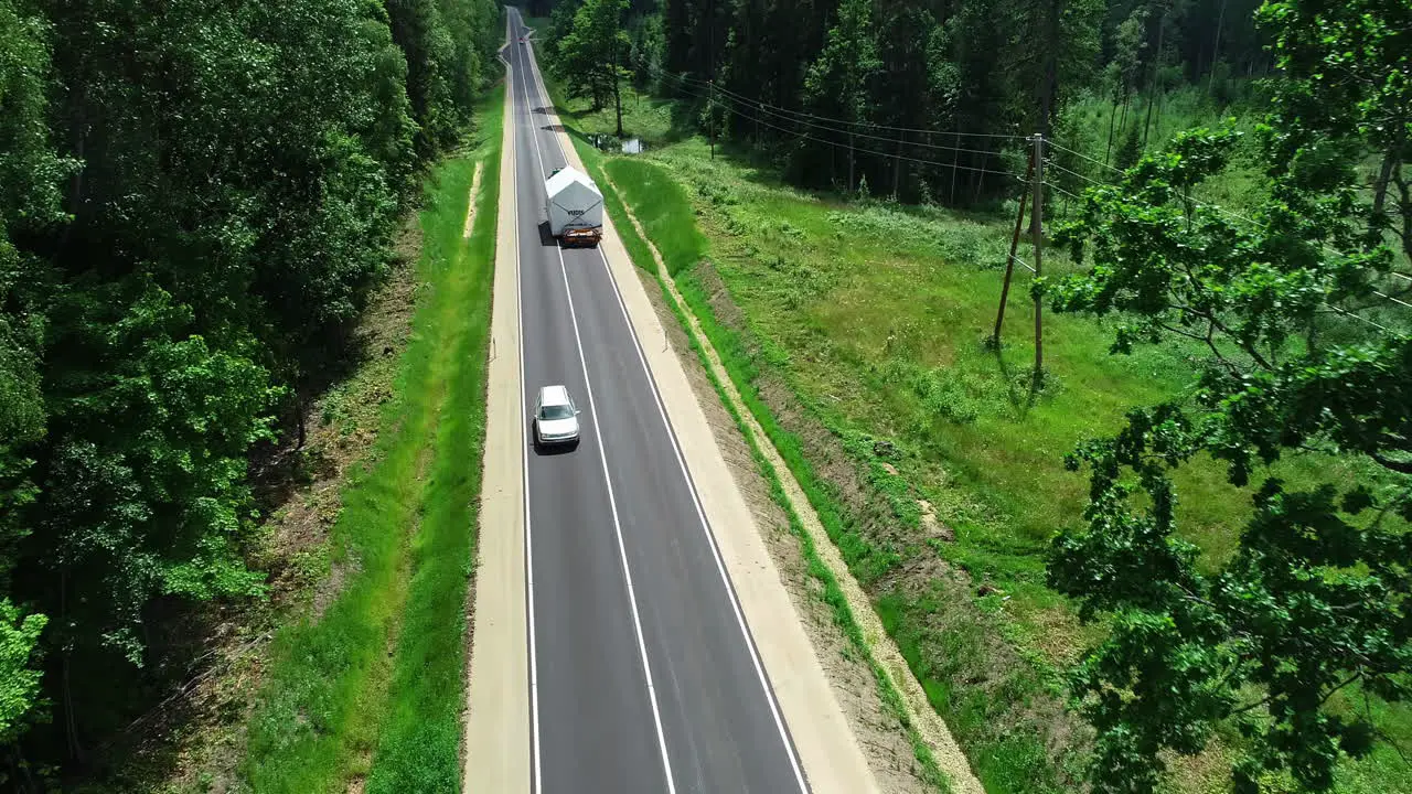 Aerial tracking shot of truck transporting prefabricated house on road between forest trees during summer