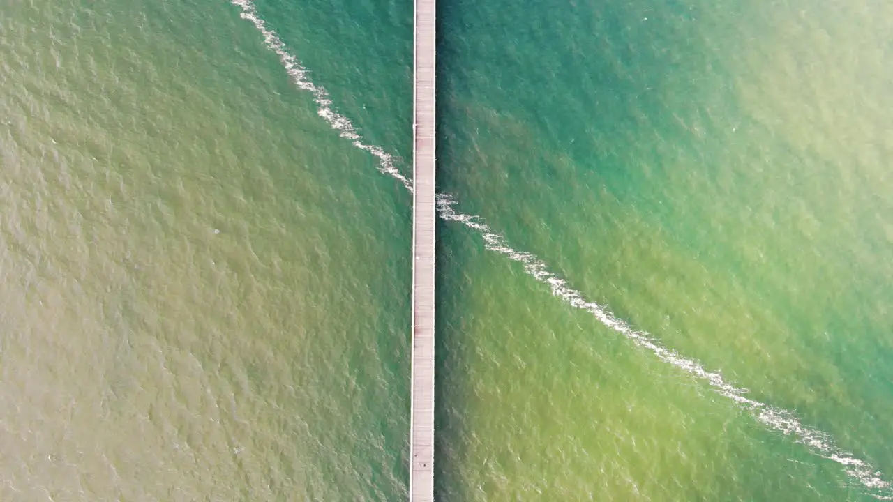 Birds eye view flyover of a long wooden pier stretching into the gorgeous clash of denim blue and lime green colours mixing in the vast ocean on a beautifully sunny day