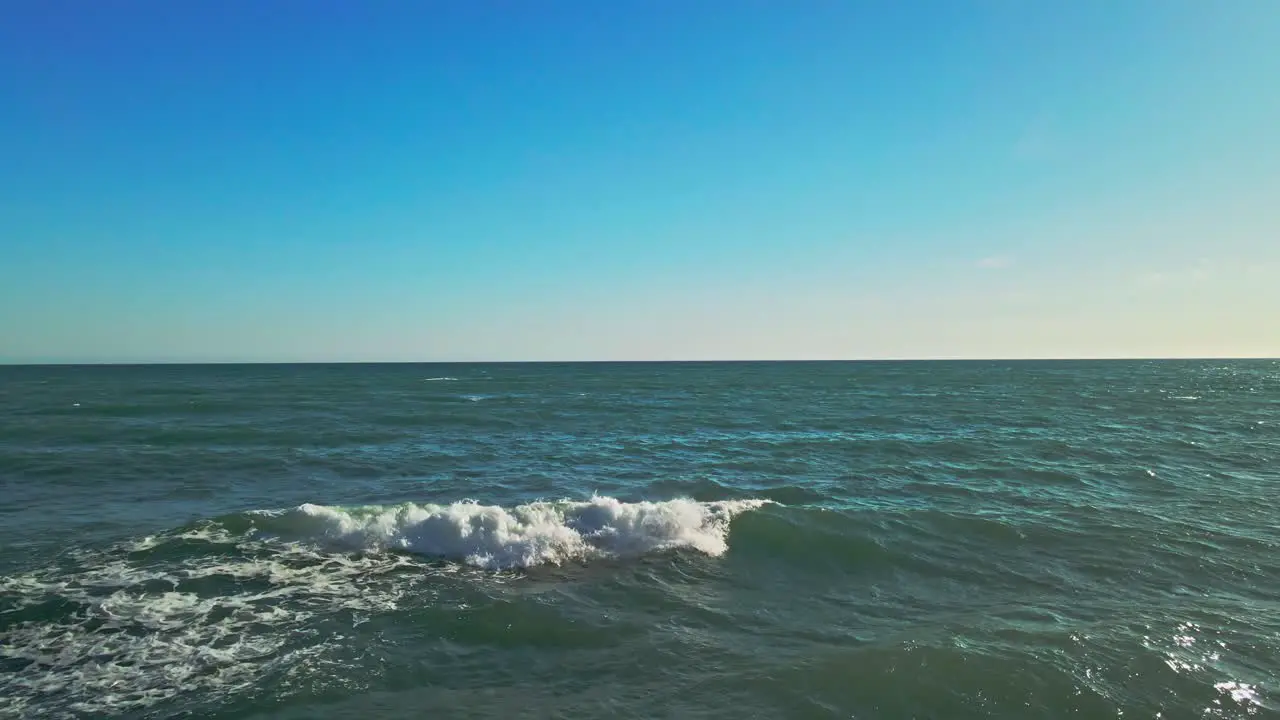 Drone in slow motion flying backwards from the sea into the rocky beach with a clear blue sky