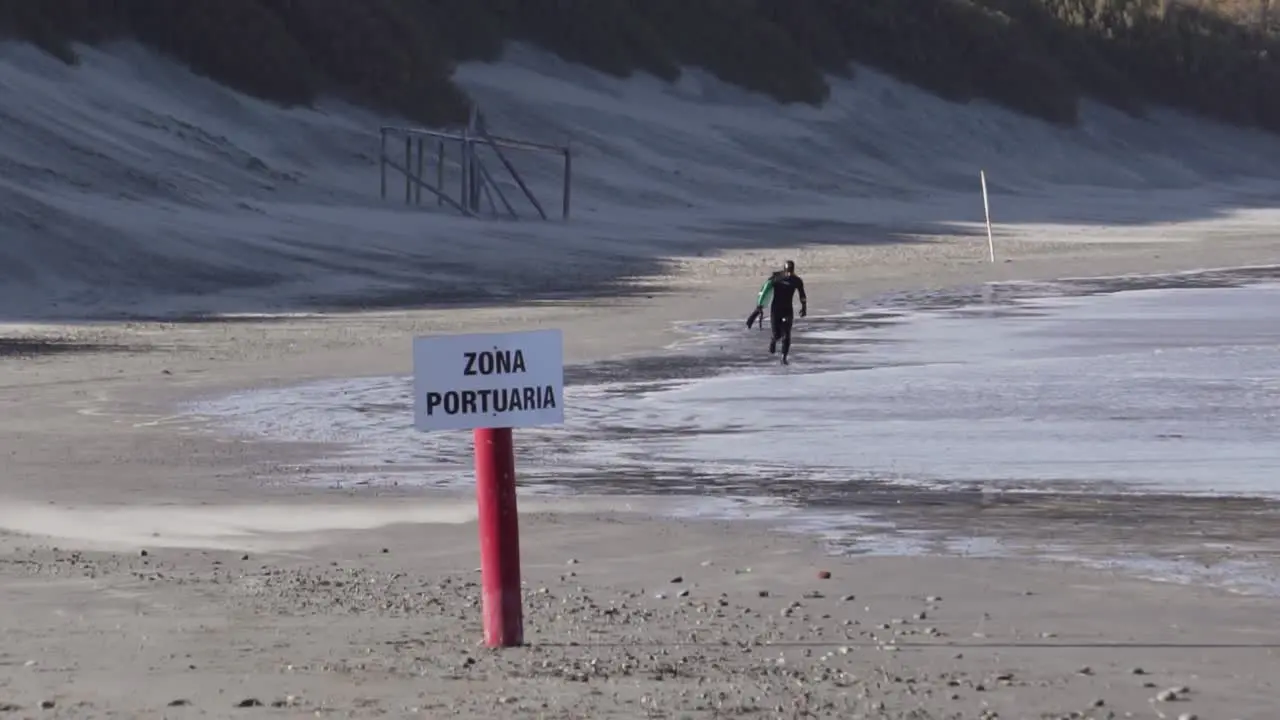 Man In Wetsuit With A Body Board Running On The Beach With Zona Portuaria Signage wide slowmo shot