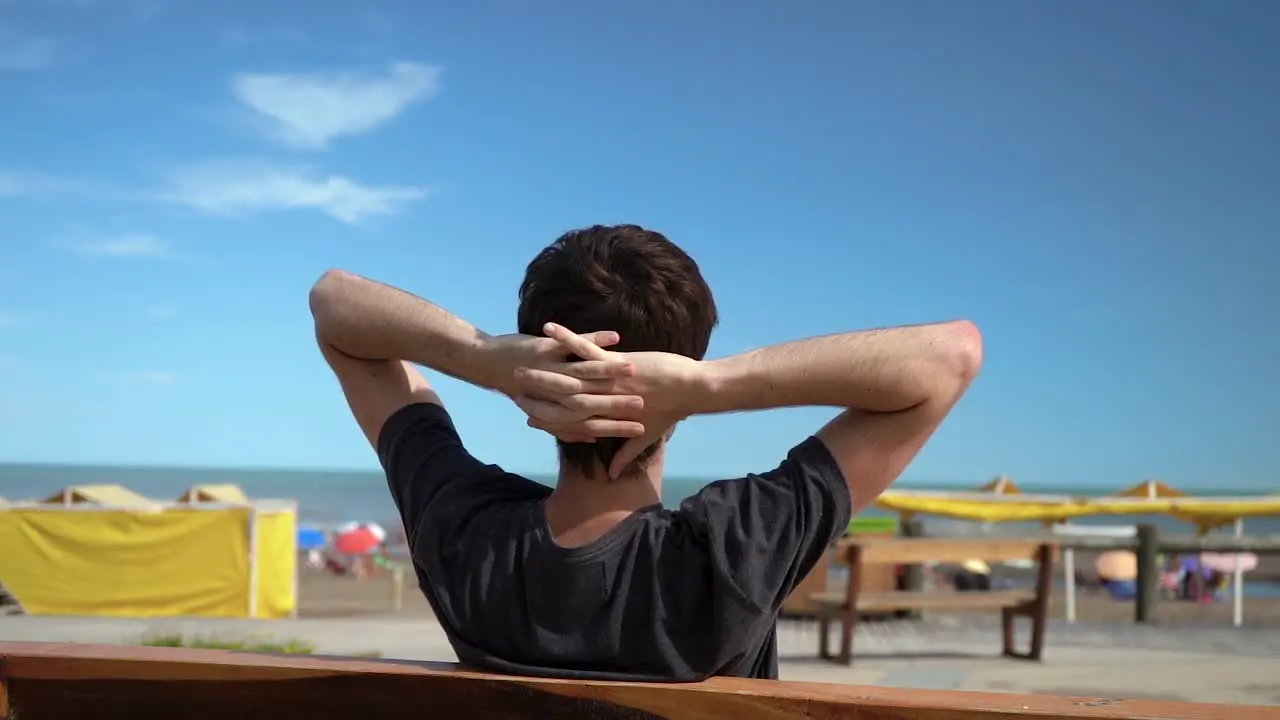 Rear Of A Man With Hands Over Back Of His Head Sitting On Wooden Bench At Summer Beachfront Of Monte Hermoso Buenos Aires Argentina