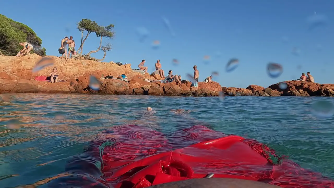 Slow motion personal perspective of man wearing red swimwear with legs and feet relaxing while floating on calm sea water with Palombaggia rocks and people in background