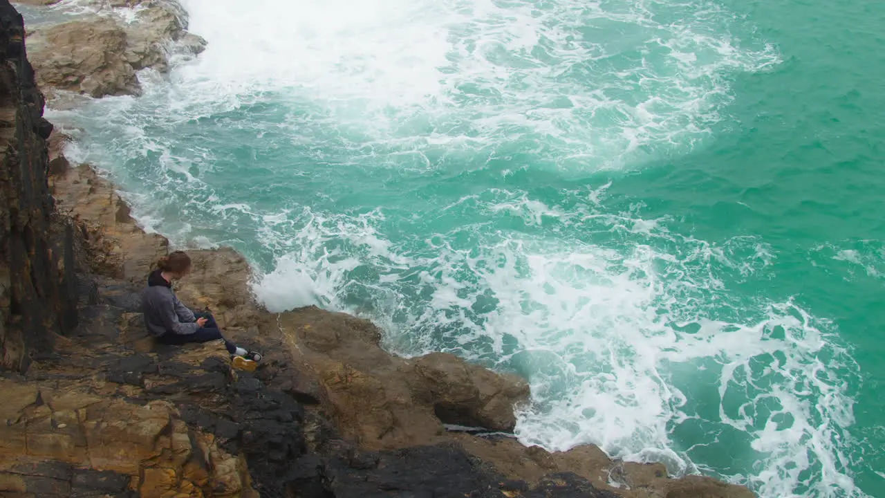 Lone Teenager Seated on Cliffside Rocks as Turquoise Ocean Waters Crash Against Rocks looking down reveal shot