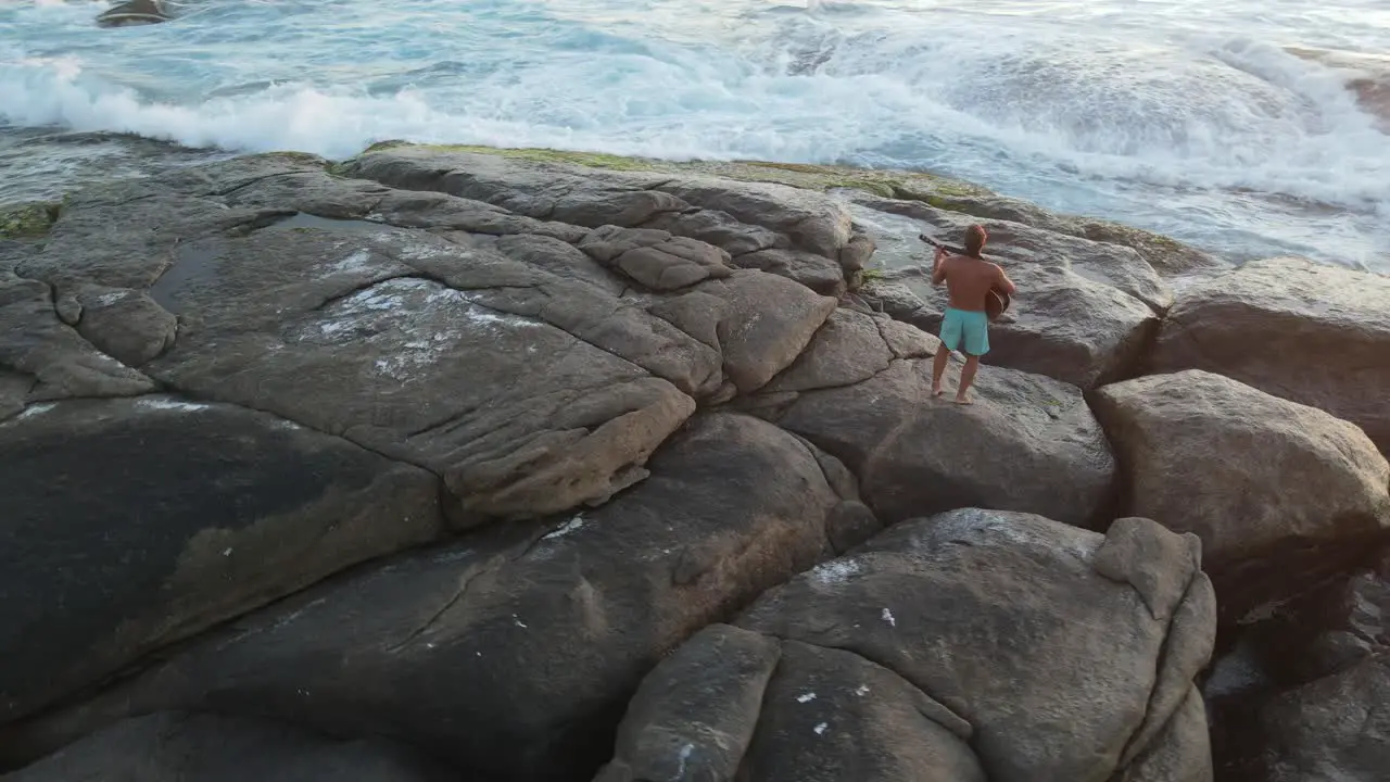 Aerial orbiting shot of young man playing emotional song on guitar during beautiful sunset at beach with crashing waves in Australia